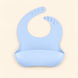 Sky Blue silicone catch bib, with a scoop pocket and adjustable button-and-hole neck closure, on warm beige.