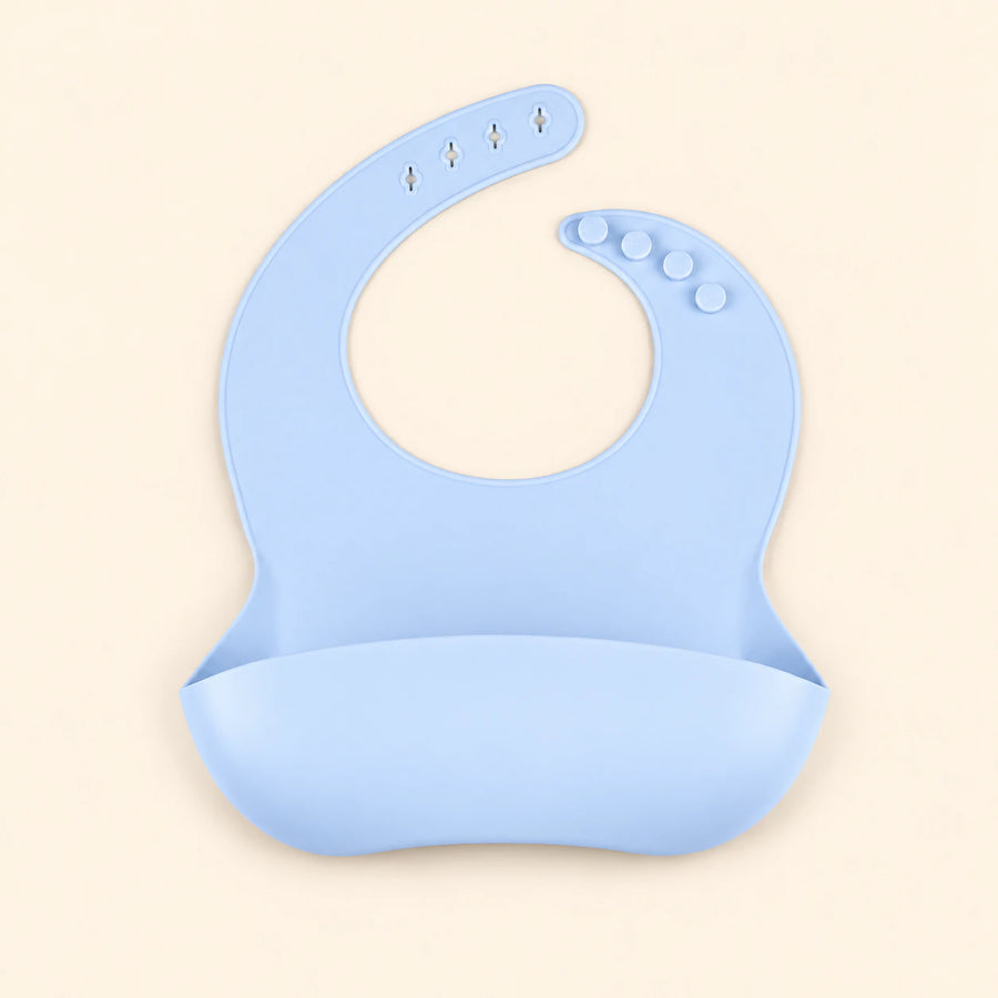 Sky Blue silicone catch bib, with a scoop pocket and adjustable button-and-hole neck closure, on warm beige.