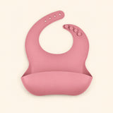 Rose Pink silicone catch bib, with a scoop pocket and adjustable button-and-hole neck closure, on warm beige.