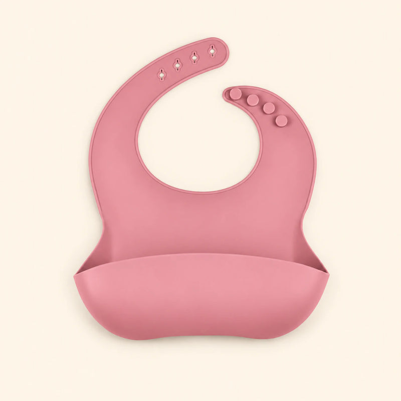 Rose Pink silicone catch bib, with a scoop pocket and adjustable button-and-hole neck closure, on warm beige.