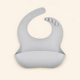 Soft Grey silicone catch bib, with a scoop pocket and adjustable button-and-hole neck closure, on warm beige.