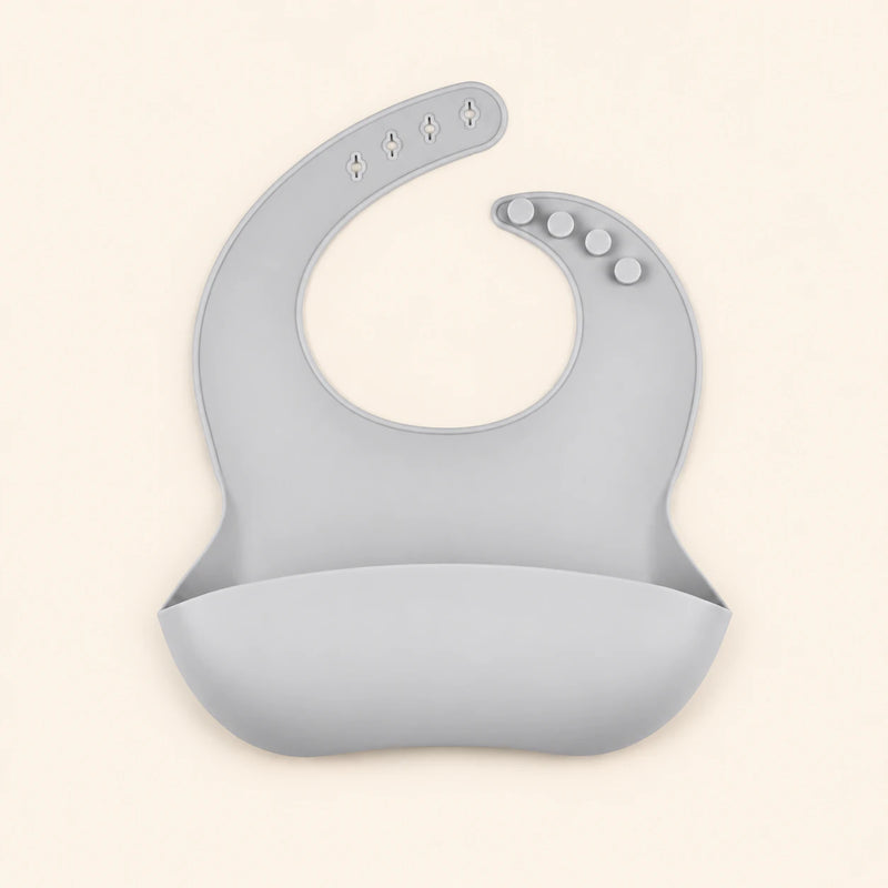 Soft Grey silicone catch bib, with a scoop pocket and adjustable button-and-hole neck closure, on warm beige.