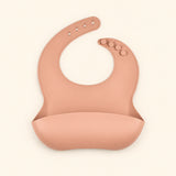 Peach silicone catch bib, with a scoop pocket and adjustable button-and-hole neck closure, on warm beige.