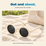 The Cream White round sunglasses rest on a beige blanket beside a canvas bag. Bag and blanket are styling props.