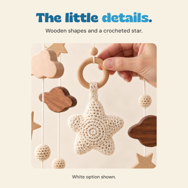 An adult hand steadies the wooden connector ring above the White mobile's complete cream crocheted star. Nearby wooden cloud shapes and cords are visible.
